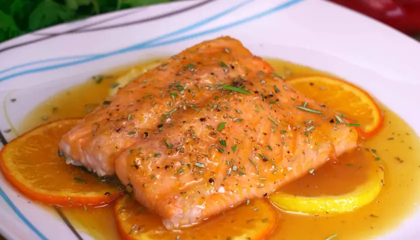 Filete de salmón a la naranja, glaseado y con hierbas, servido sobre rodajas de naranja y limón en una salsa dorada.