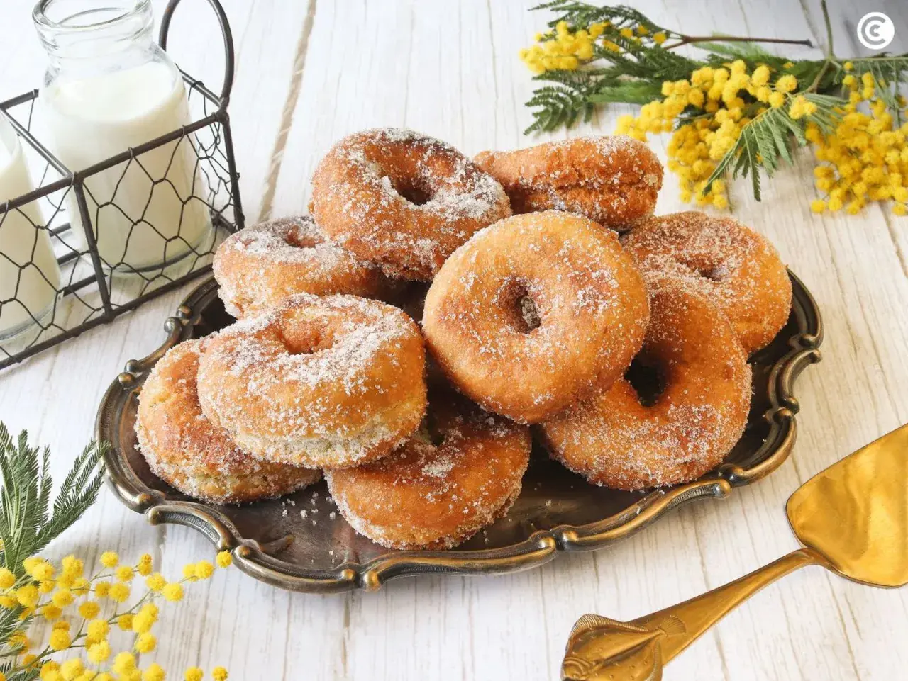 Un plato de rosquillas caseras de toda la vida, espolvoreadas con azúcar, junto a botellas de leche y flores amarillas.