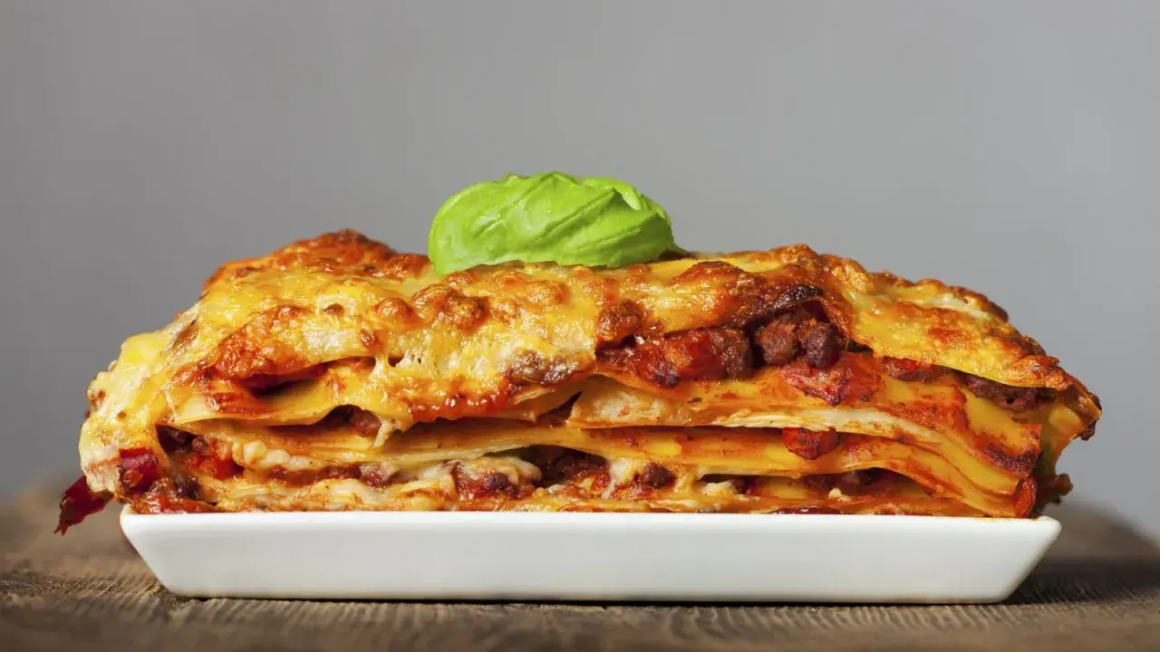 Una porción generosa de lasaña de carne, con capas de pasta, salsa y queso dorado, coronada con una hoja de albahaca fresca.