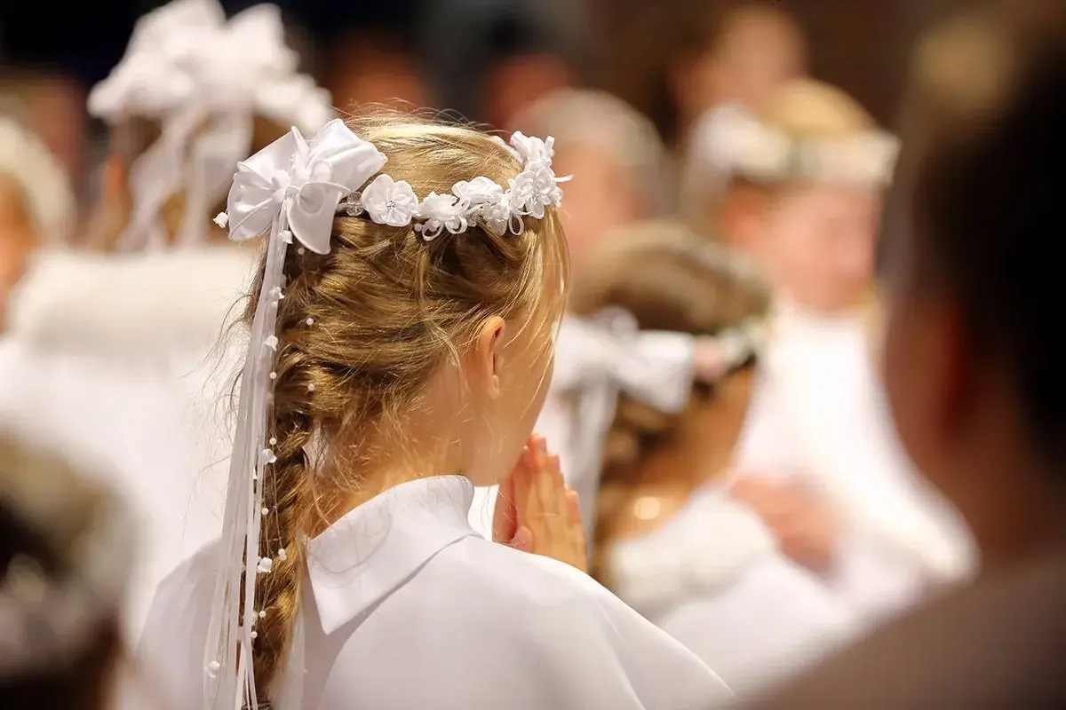 Niña con corona de flores y trenza adornada, rezando en una ceremonia de comunión.