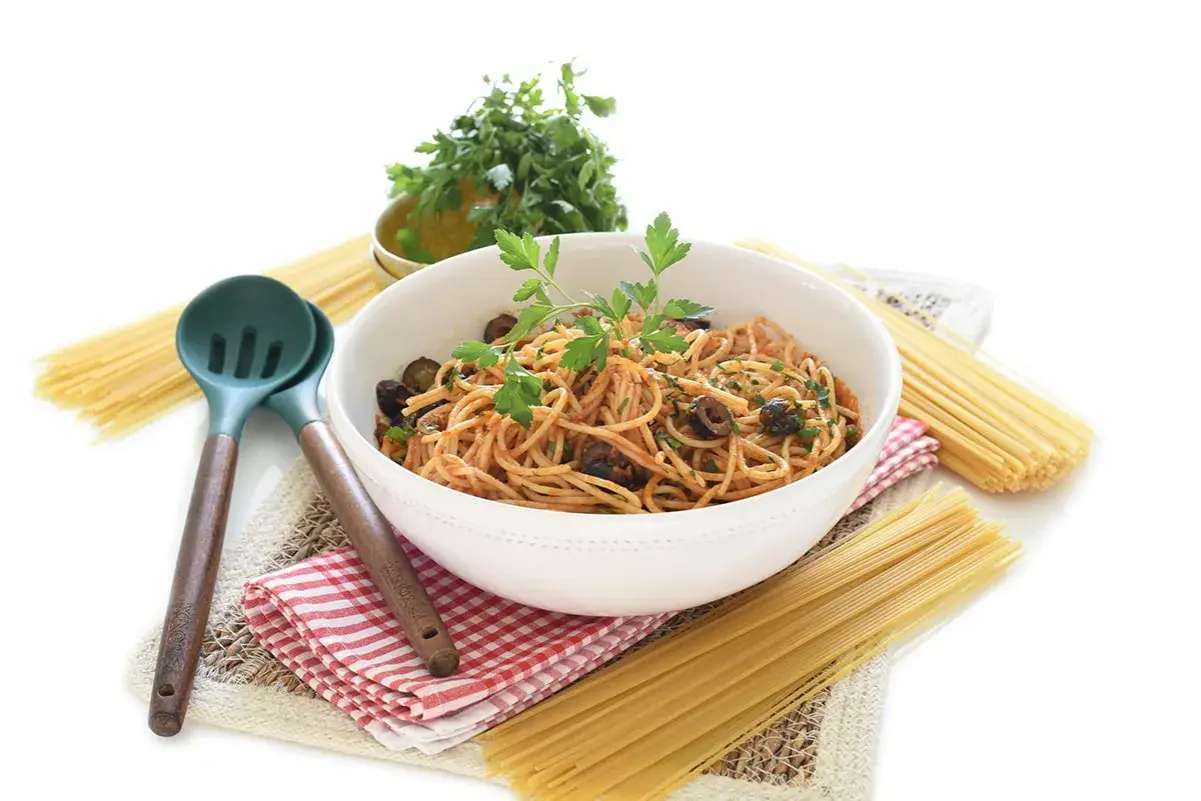 Un tazón de espagueti con salsa putanesca, aceitunas negras y perejil fresco, acompañado de pasta cruda y utensilios de cocina.