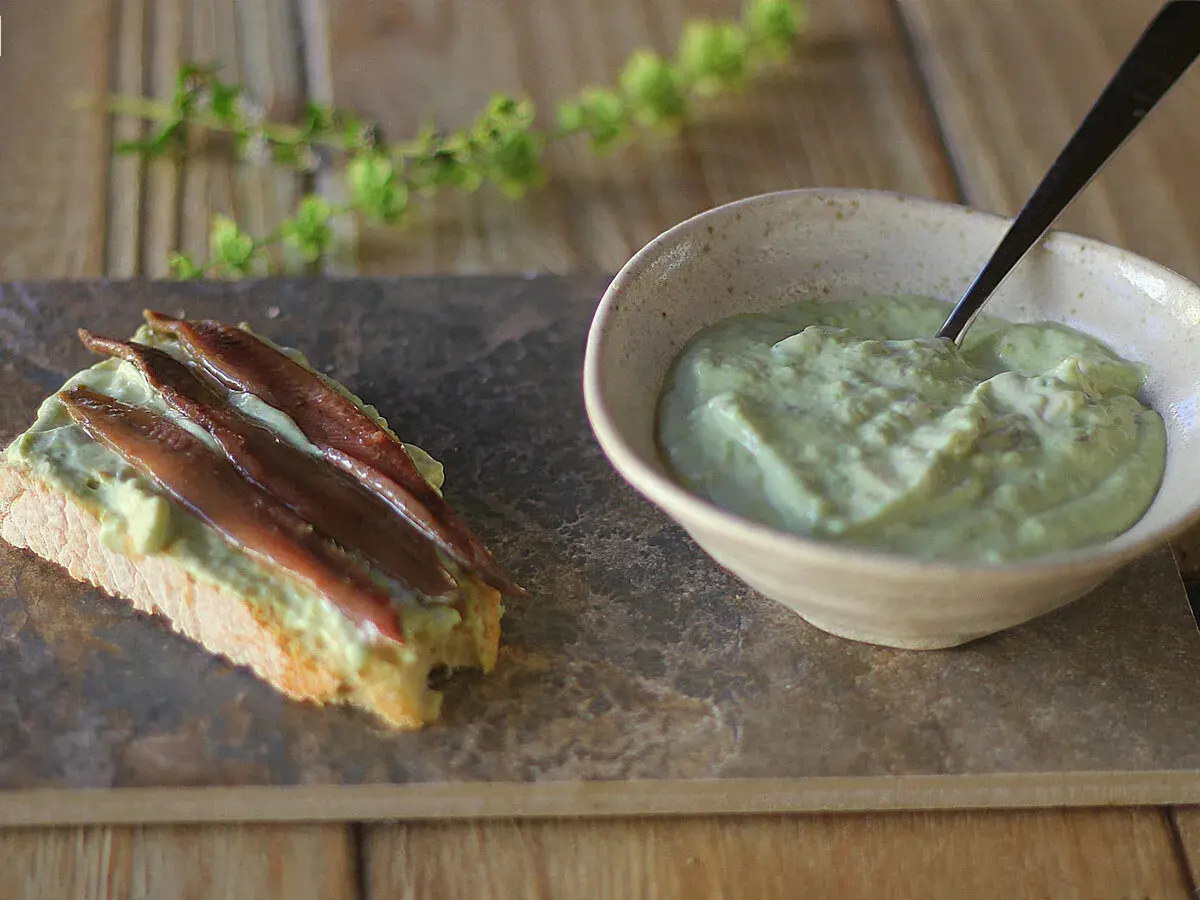 Tosta con mayonesa de aguacate y anchoas. Un cuenco con la cremosa mayonesa de aguacate y una tostada decorada con ella y filetes de anchoa.