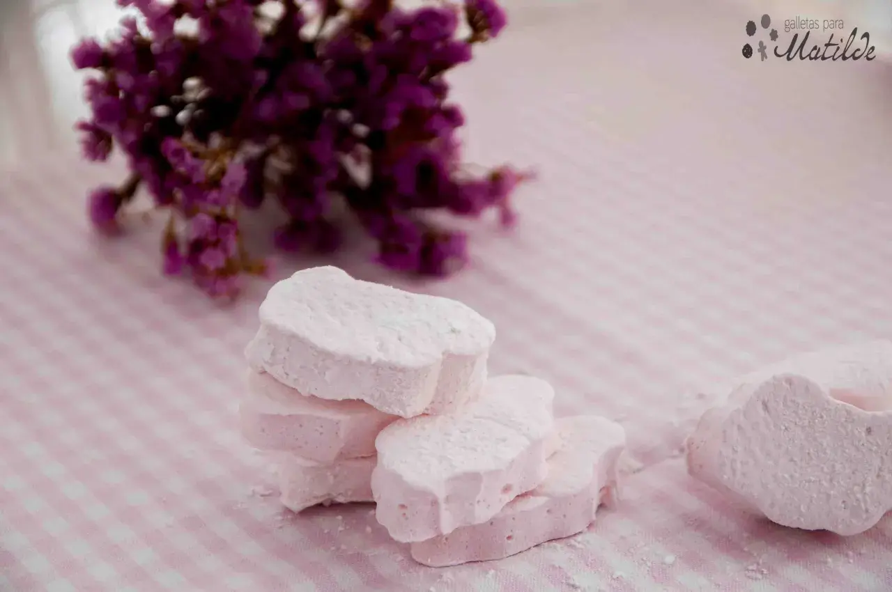 Delicados nubes dulces rosados, espolvoreados con azúcar glas, junto a un ramo de flores moradas sobre un mantel de cuadros.