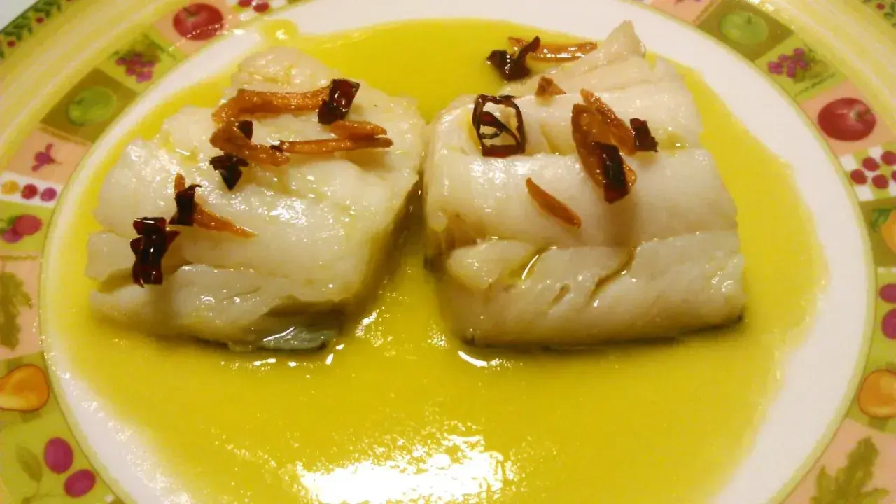 Dos trozos de bacalao al pil pil, cubiertos con aceite de oliva y adornados con ajo frito y guindilla.