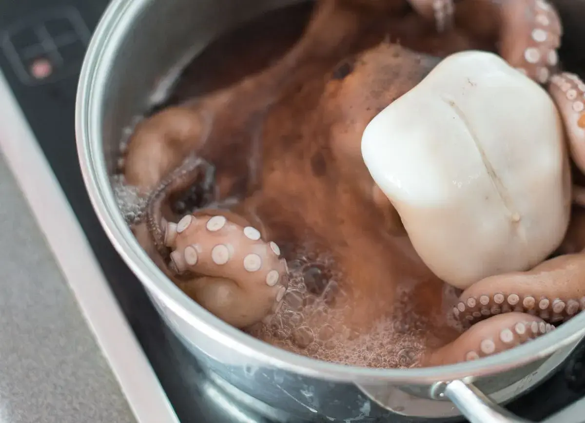 Un pulpo entero se está cociendo en una olla de metal. Sus tentáculos rosados con ventosas visibles se curvan en el agua burbujeante.