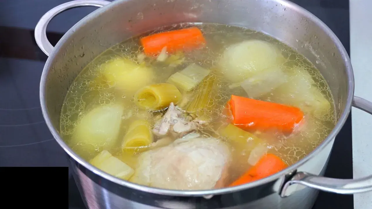 Olla con caldo de pollo casero burbujeante, con trozos de zanahoria, puerro y patata.