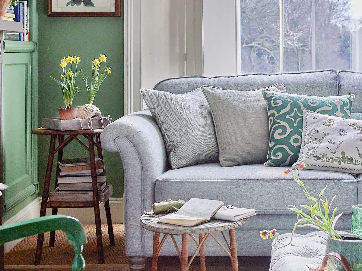 Sofá azul claro con cojines decorativos. Una mesa auxiliar con narcisos y libros complementa la decoración color pastel.