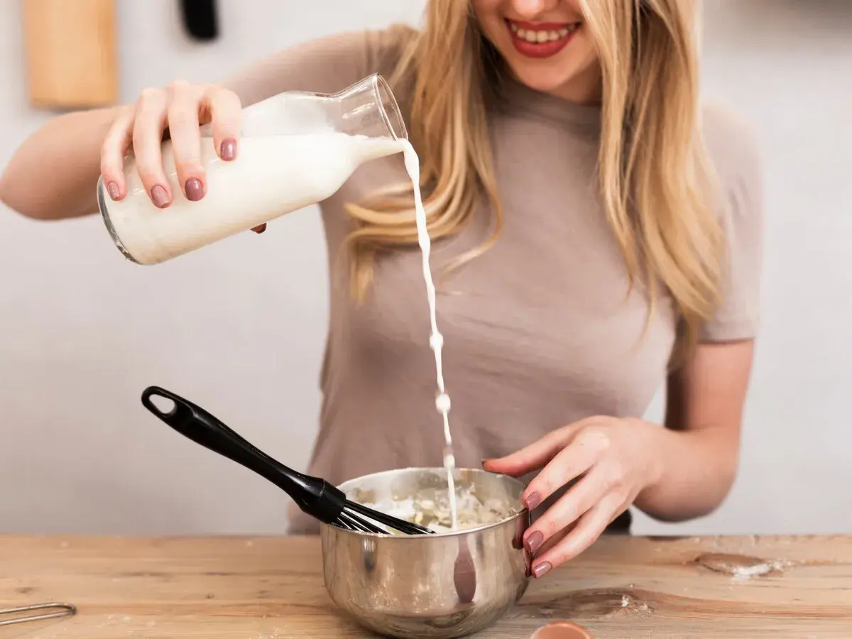 Mujer rubia sonríe mientras vierte leche de una botella a un bol con batidor, preparando algo delicioso.