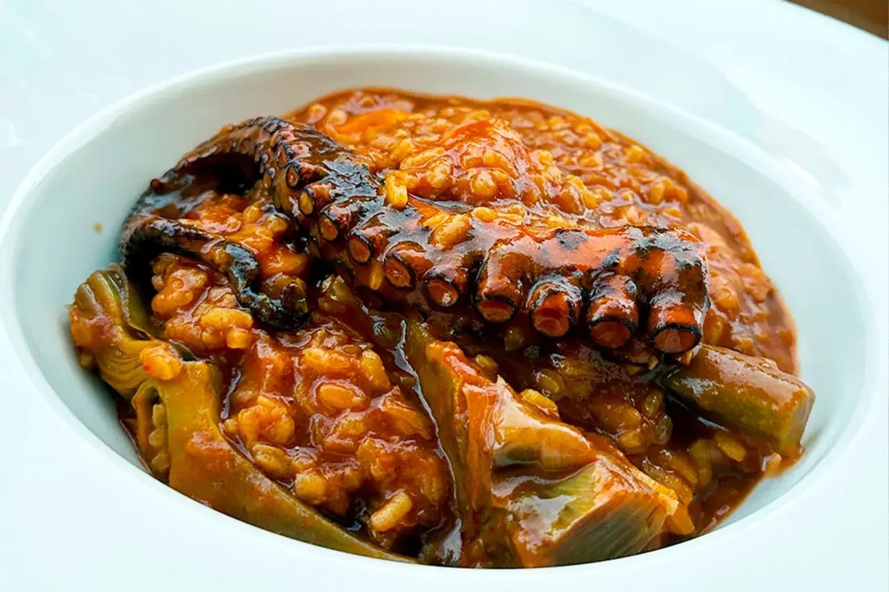 Un tentáculo de pulpo asado reposa sobre un arroz meloso y sabroso, con trozos de alcachofa. Un delicioso arroz con pulpo.