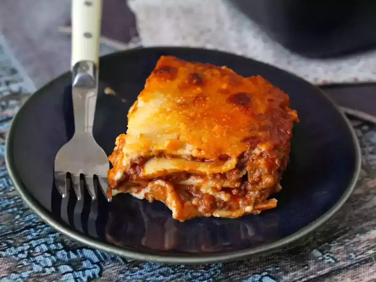 Un trozo de lasaña boloñesa, con capas de pasta, carne y salsa, servido en un plato azul con un tenedor.