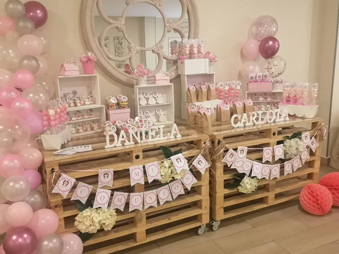 Mesa dulce para comunión de niña, con candy bar casero, globos rosas y blancos, y detalles personalizados.