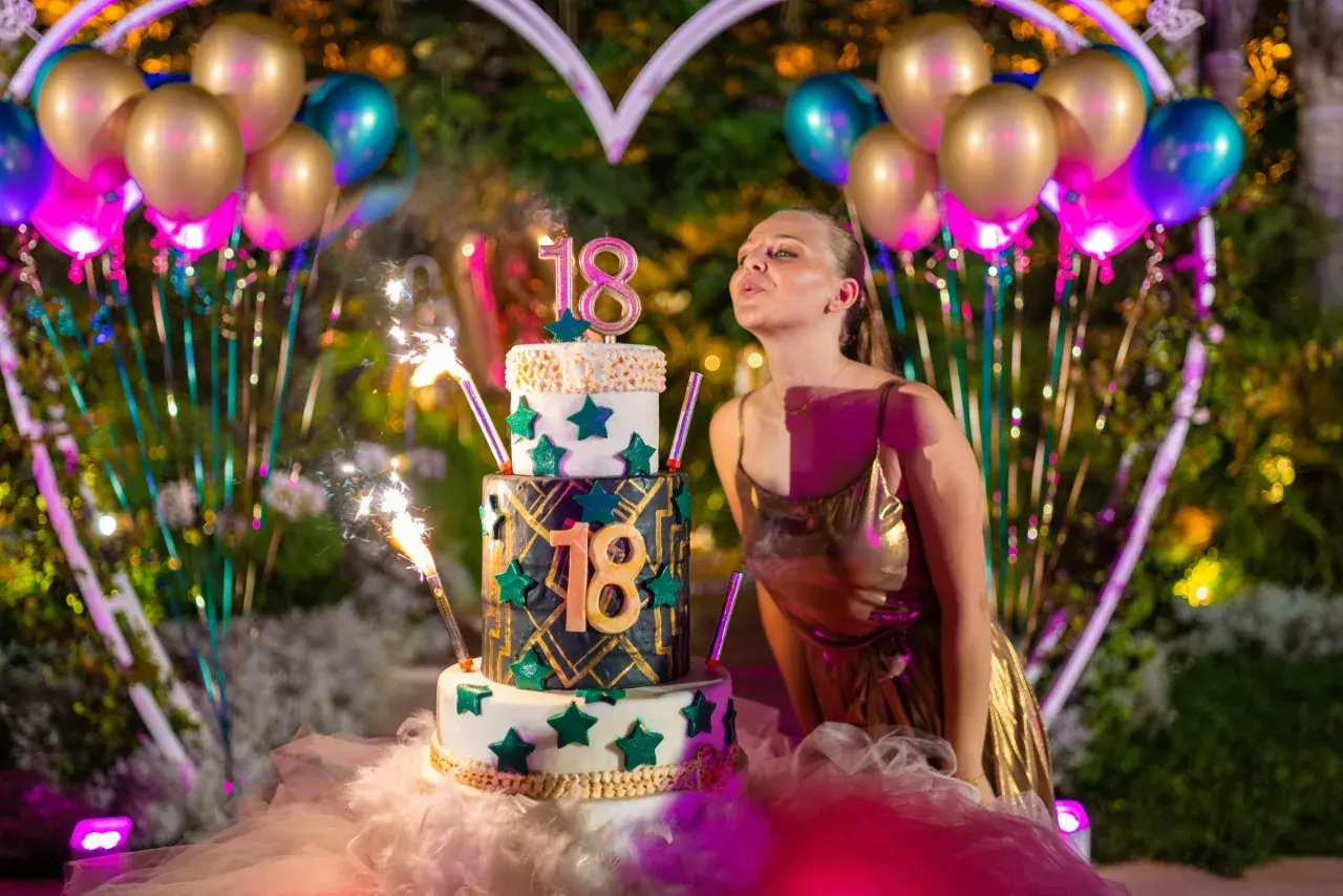 Joven soplando velas en un pastel de tres pisos para celebrar su 18 cumpleaños, rodeada de globos y luces.