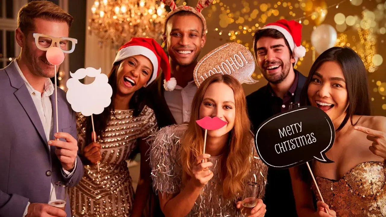 Grupo de amigos sonriendo y riendo en un photocall navideño casero, con accesorios como gafas, barba, labios y carteles de "Ho ho ho" y "Merry Christmas".