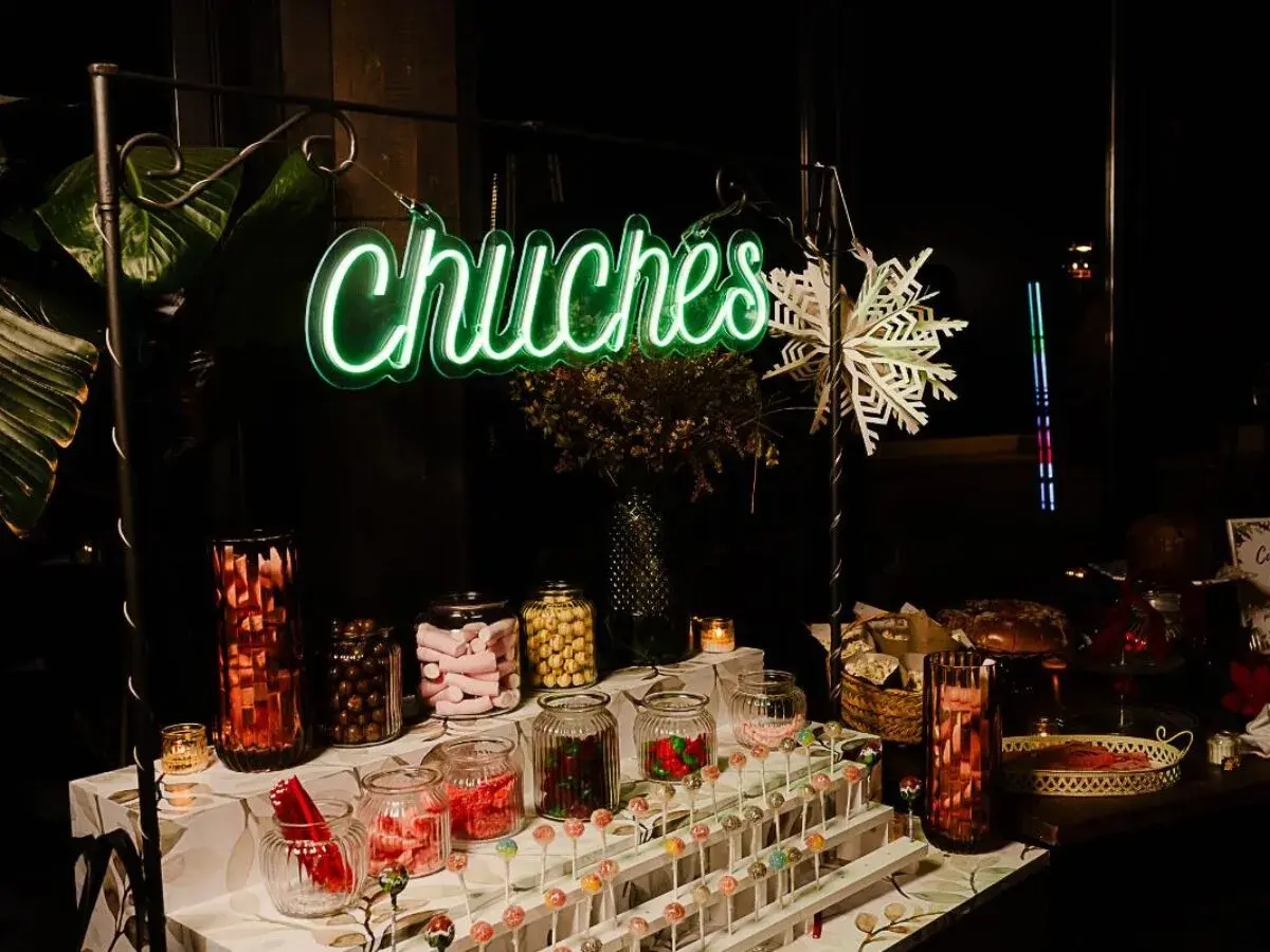 Un letrero de neón verde "Chuchas" ilumina un candy bar lleno de frascos de dulces, piruletas y golosinas.