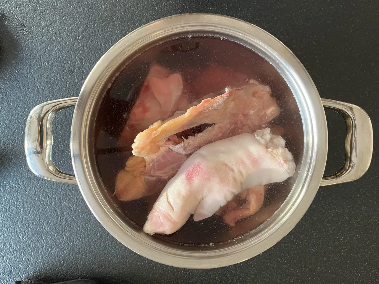 Olla con ingredientes para caldo de huesos: patas de cerdo, huesos y trozos de carne en un líquido oscuro.