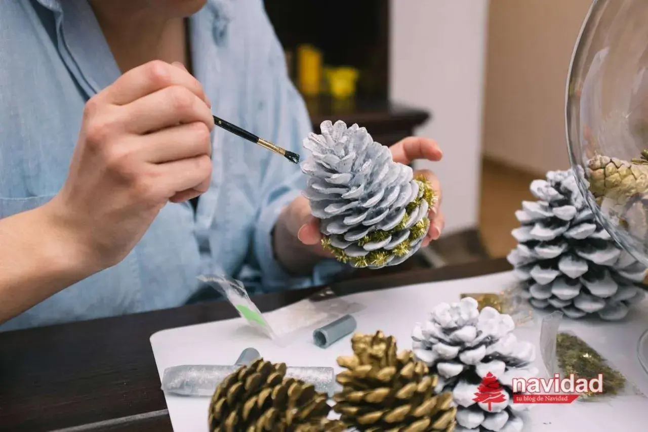 Manos pintando piñas para navidad con pintura plateada y detalles dorados. Decoración festiva en proceso.