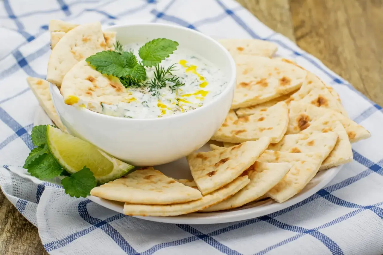 Un cuenco de cremosa salsa tzatziki, decorado con hierbas frescas y un chorrito de aceite, servido con pan de pita crujiente y rodajas de lima.
