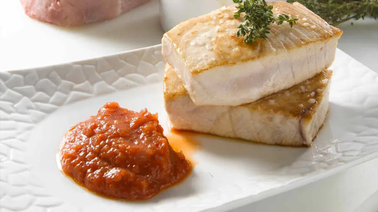 Dos trozos de atún a la plancha, dorados y jugosos, apilados sobre un plato blanco con una porción de salsa de tomate y una ramita de tomillo.