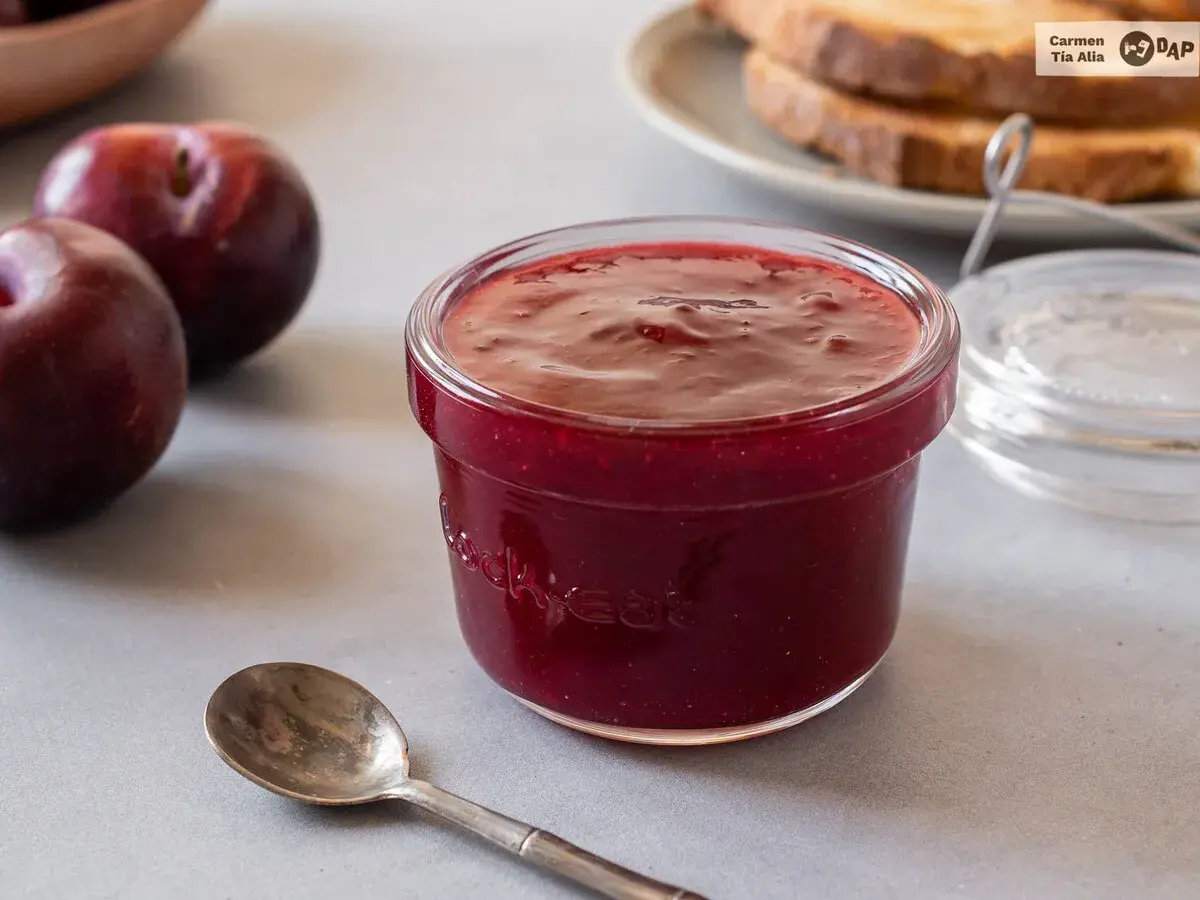 Un tarro de mermelada de ciruela casera, con ciruelas frescas y tostadas al fondo. ¡Perfecta para el desayuno!