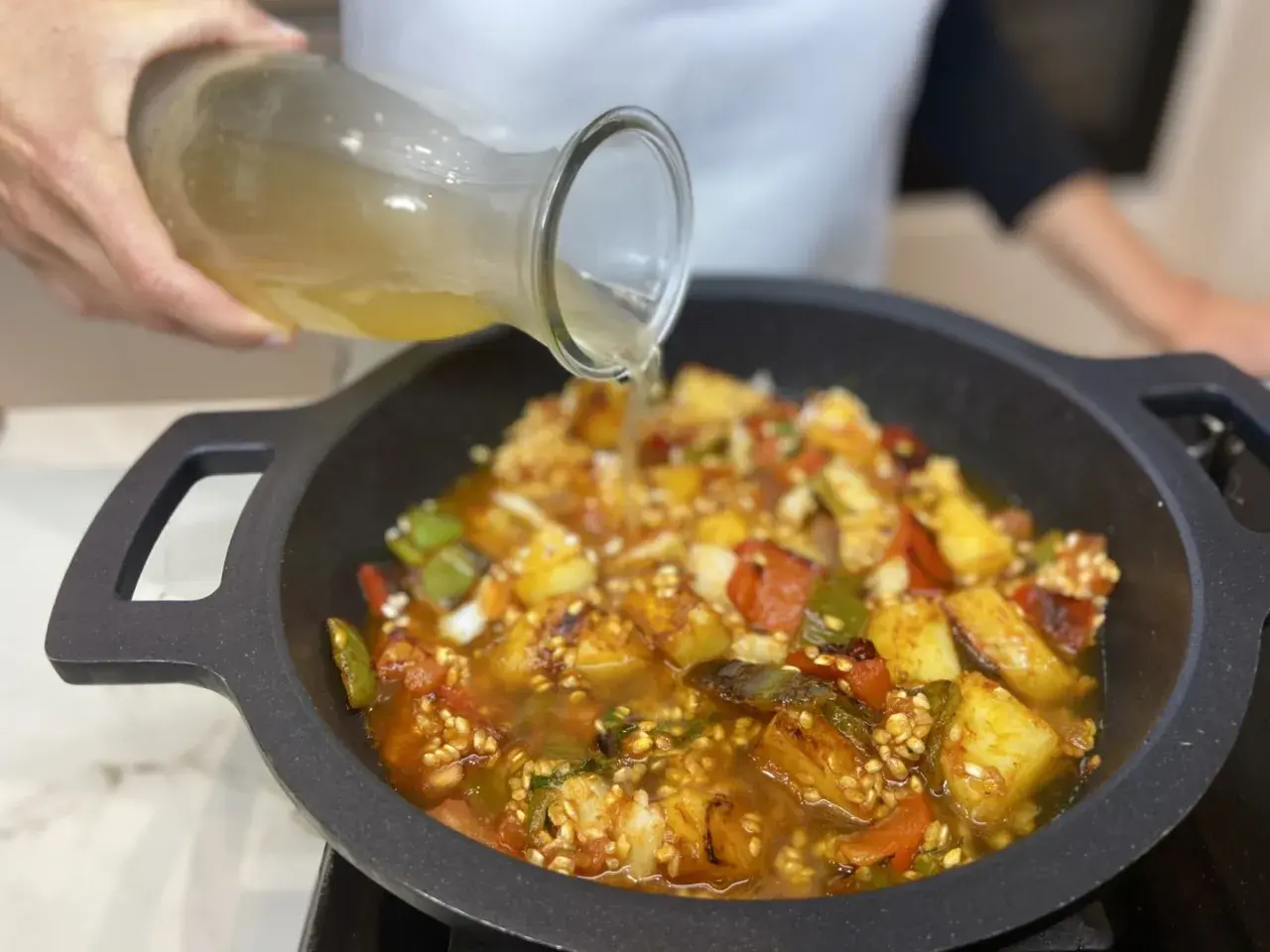 Se vierte caldo sobre un sofrito para paella con pimientos, tomate y patatas en una paellera.