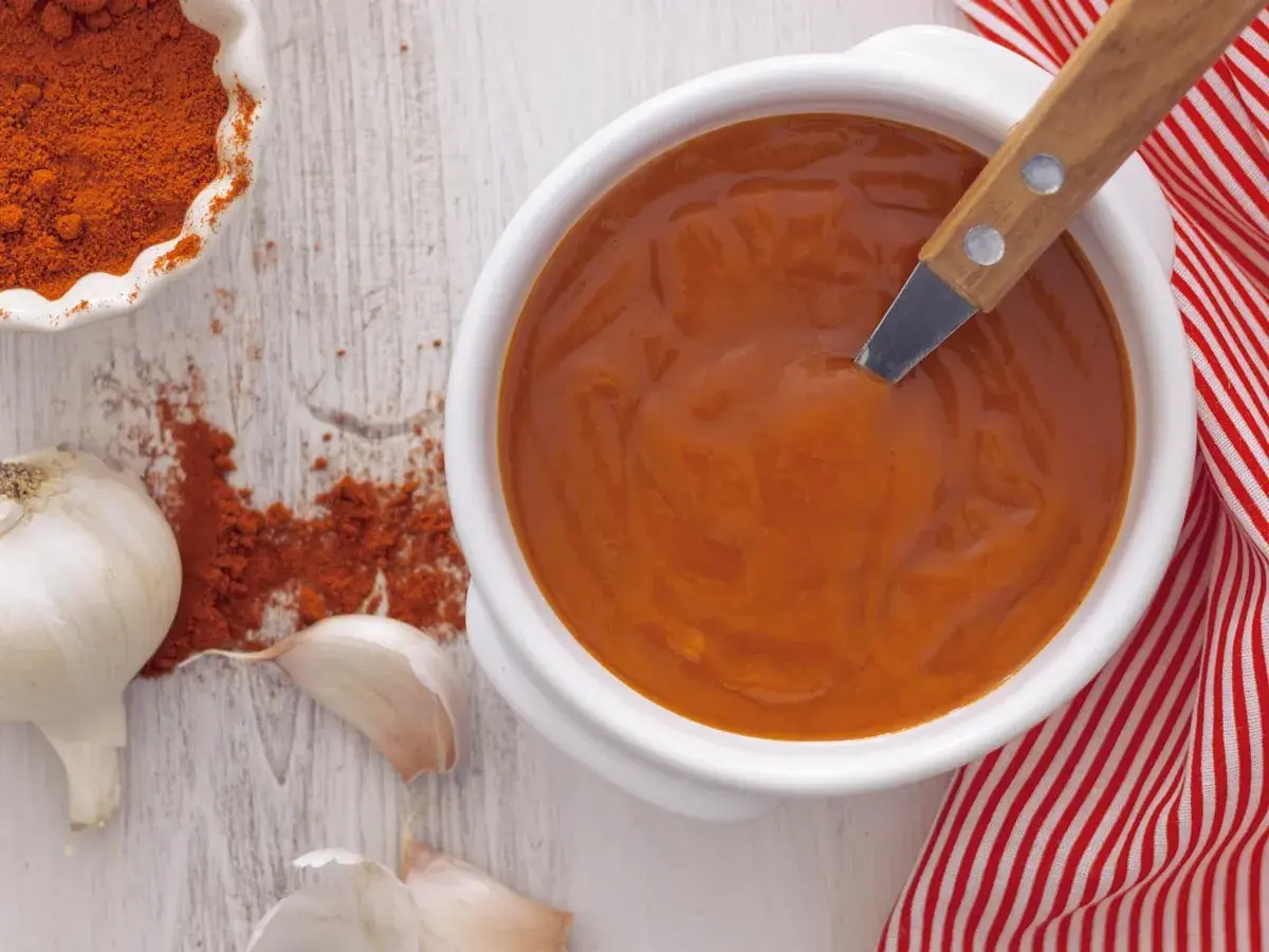 Un cuenco blanco con salsa brava, pimentón en polvo, ajos y una servilleta a rayas rojas.