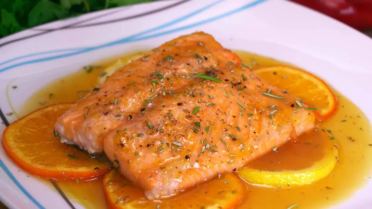 Filete de salmón a la naranja, glaseado y con hierbas, servido sobre rodajas de naranja y limón en una salsa dorada.