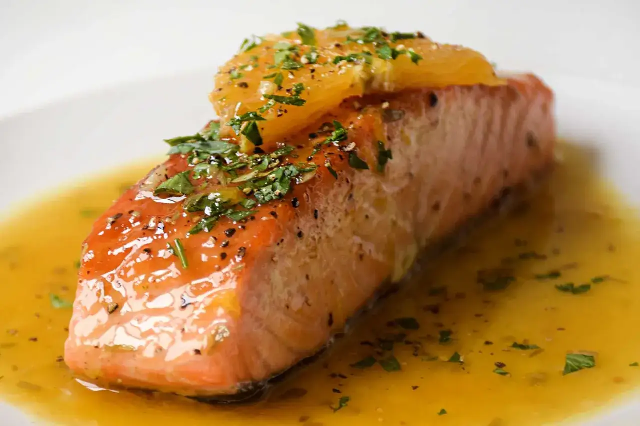 Delicioso salmón a la naranja, bañado en su jugo y adornado con hierbas frescas y una rodaja de naranja.