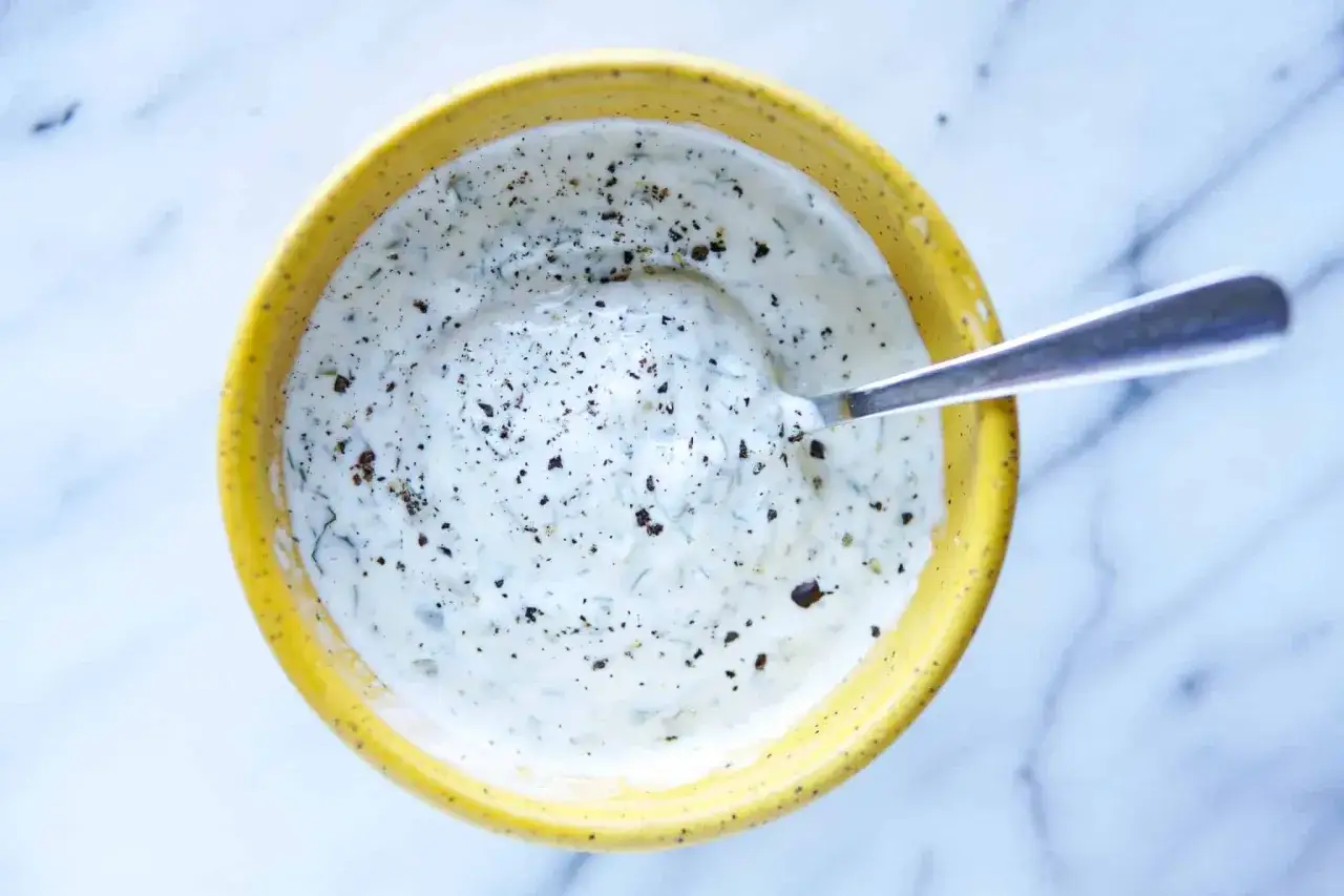 Un cuenco amarillo contiene una cremosa salsa de yogur con hierbas y pimienta negra. Una cuchara plateada reposa en ella.
