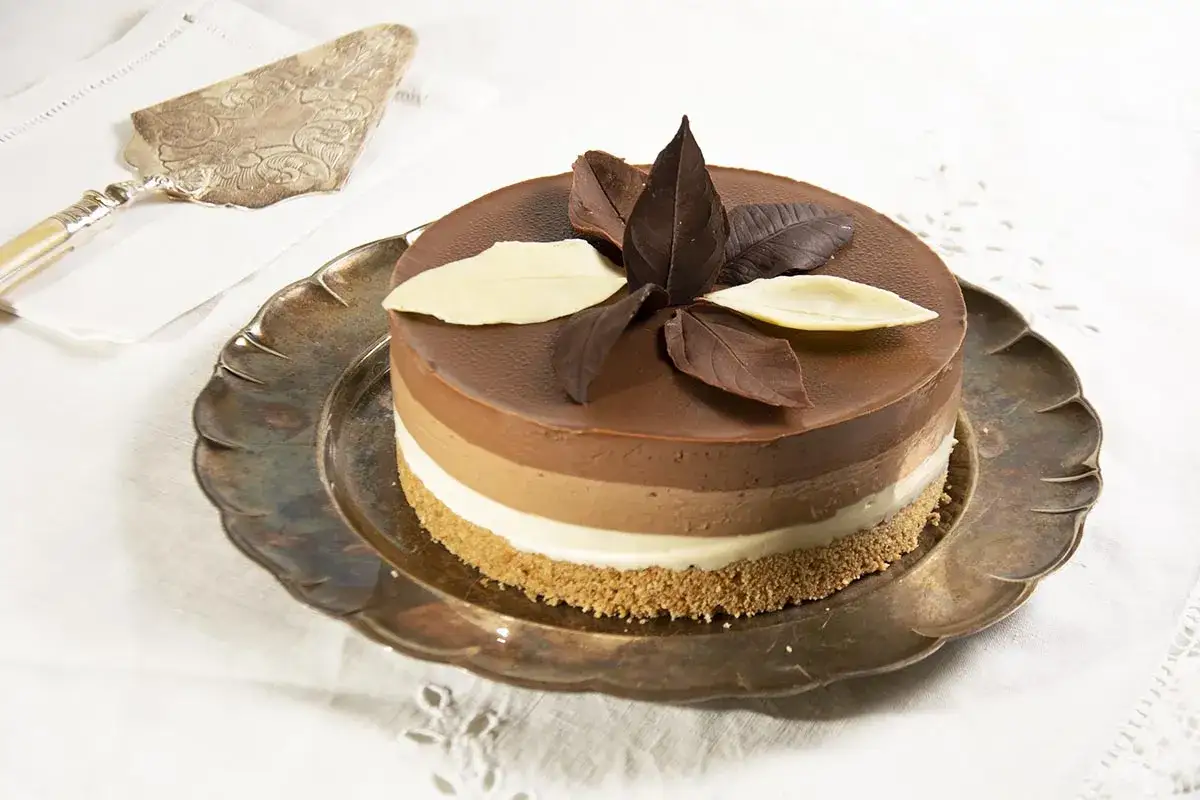 Deliciosa tarta de tres chocolates, con capas de mousse de chocolate blanco, con leche y negro, decorada con hojas de chocolate.