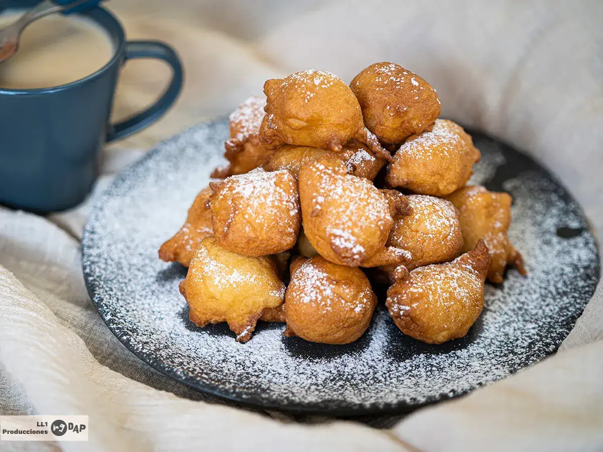 Mont&oacute;n de bu&ntilde;uelos de manzana dorados y espolvoreados con az&uacute;car glas, junto a una taza de caf&eacute;.