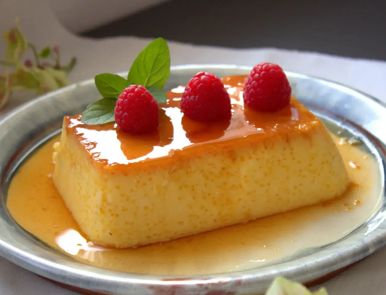 Un trozo de flan de queso cremoso, bañado en caramelo y adornado con frambuesas y menta.