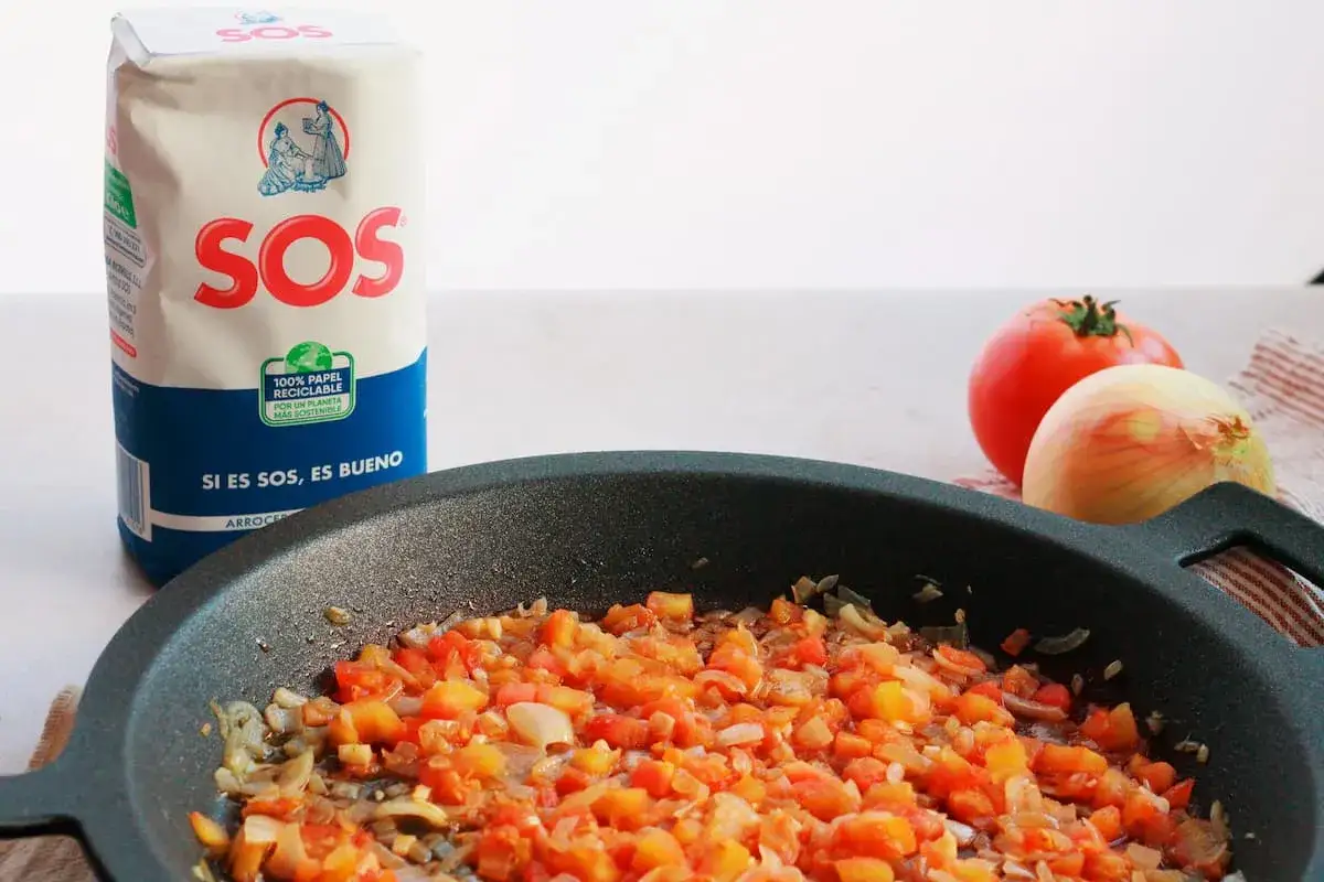 Sofrito para paella en sartén, con tomate y cebolla picados. Bolsa de arroz SOS al fondo.