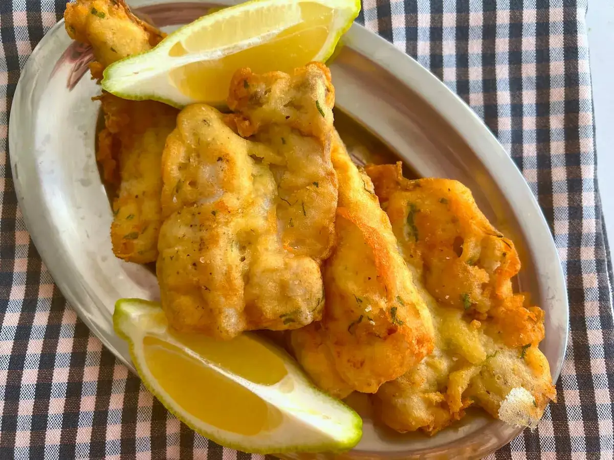 Crujiente merluza a la romana, dorada y servida con gajos de lim&oacute;n fresco en una bandeja met&aacute;lica.
