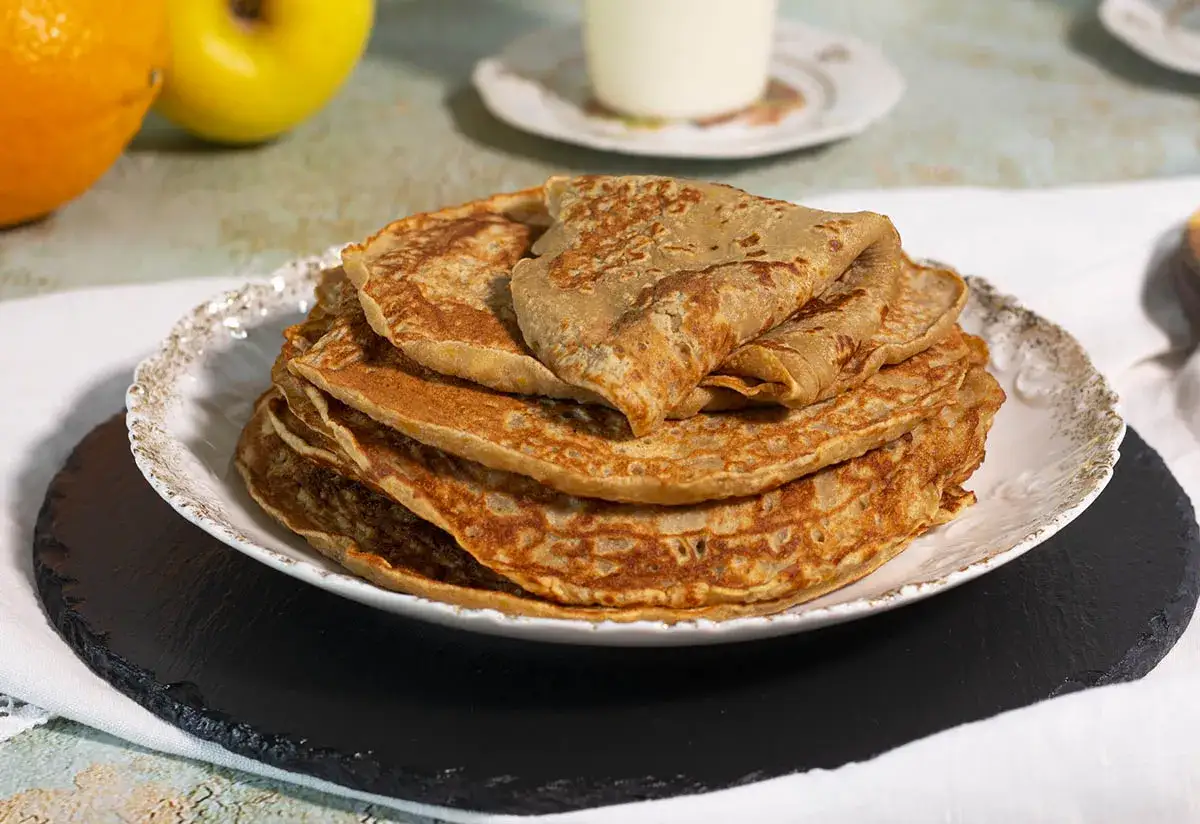 Pila de tortitas de avena doradas y esponjosas en un plato blanco con borde dorado.