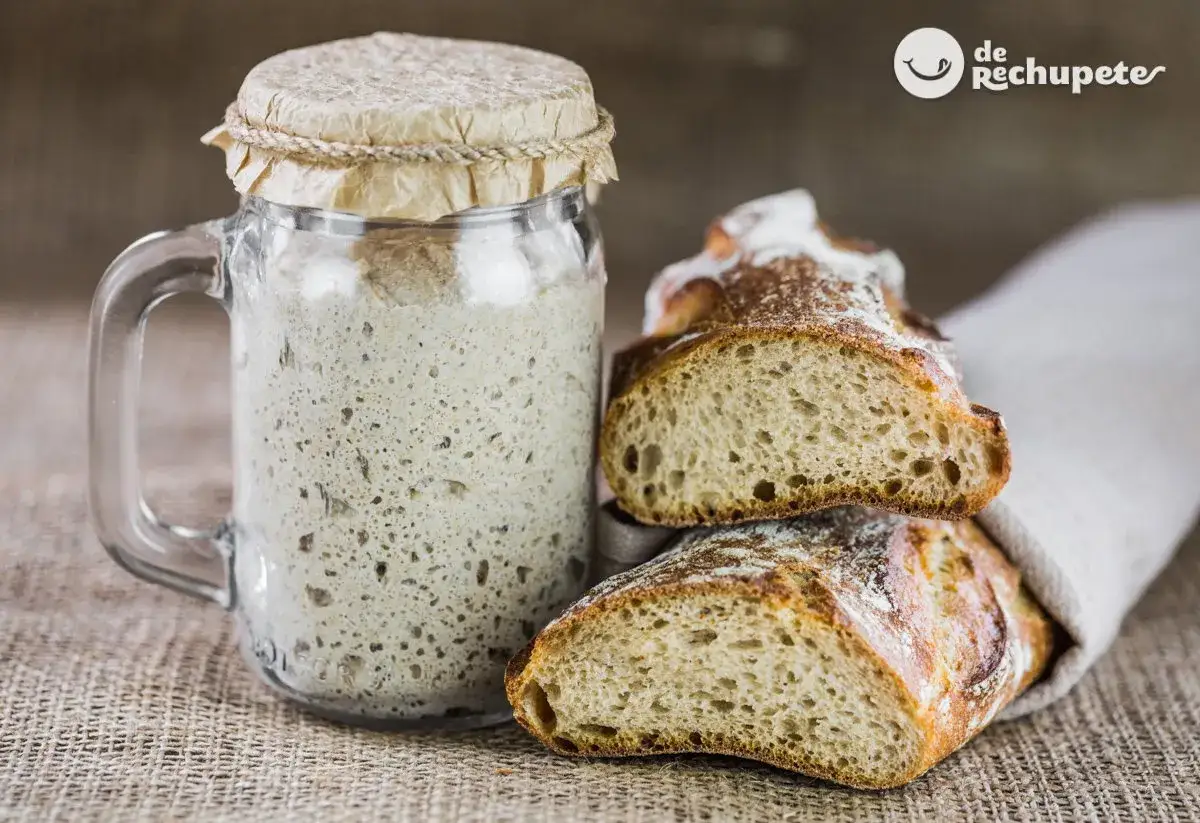 Tarro con masa madre burbujeante junto a rebanadas de pan artesanal, listo para hornear.