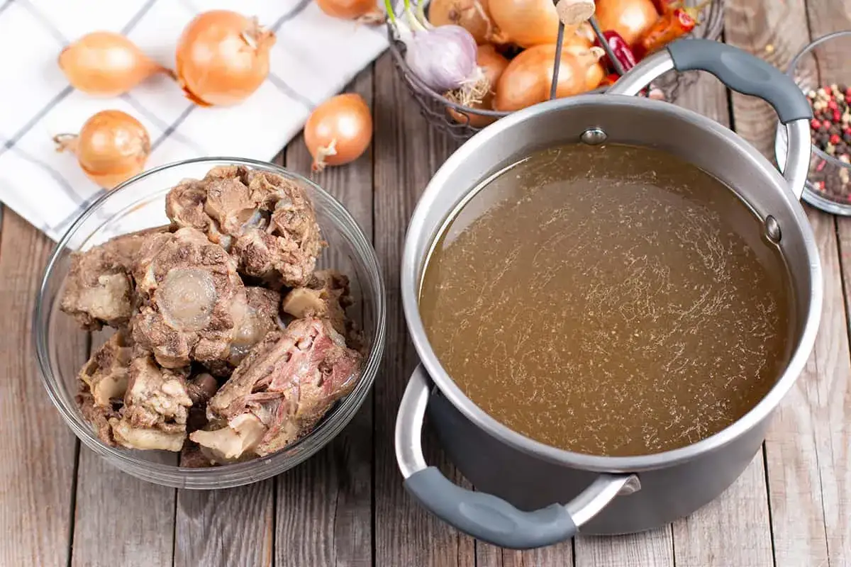 Olla con caldo de huesos, junto a un bol con trozos de rabo de toro y cebollas.