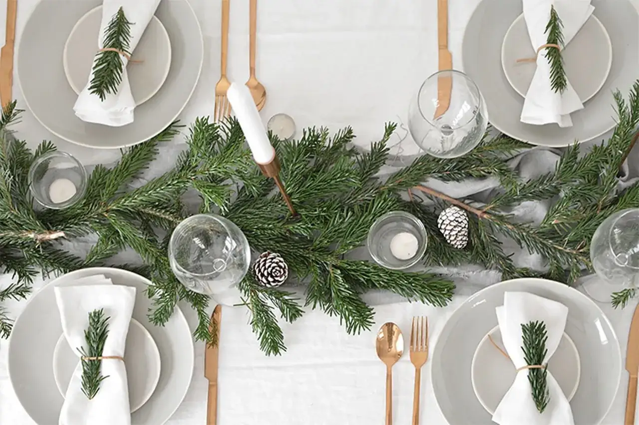 Elegantes mesas decoradas de navidad con guirnaldas de pino, pi&ntilde;as y velas. Vajilla gris, servilletas blancas con ramitas de abeto y cuberter&iacute;a dorada.