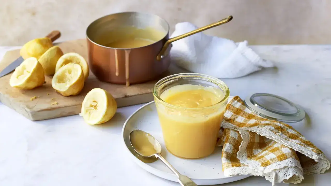 Un tarro de cremoso lemon curd, junto a limones exprimidos y una cacerola de cobre.