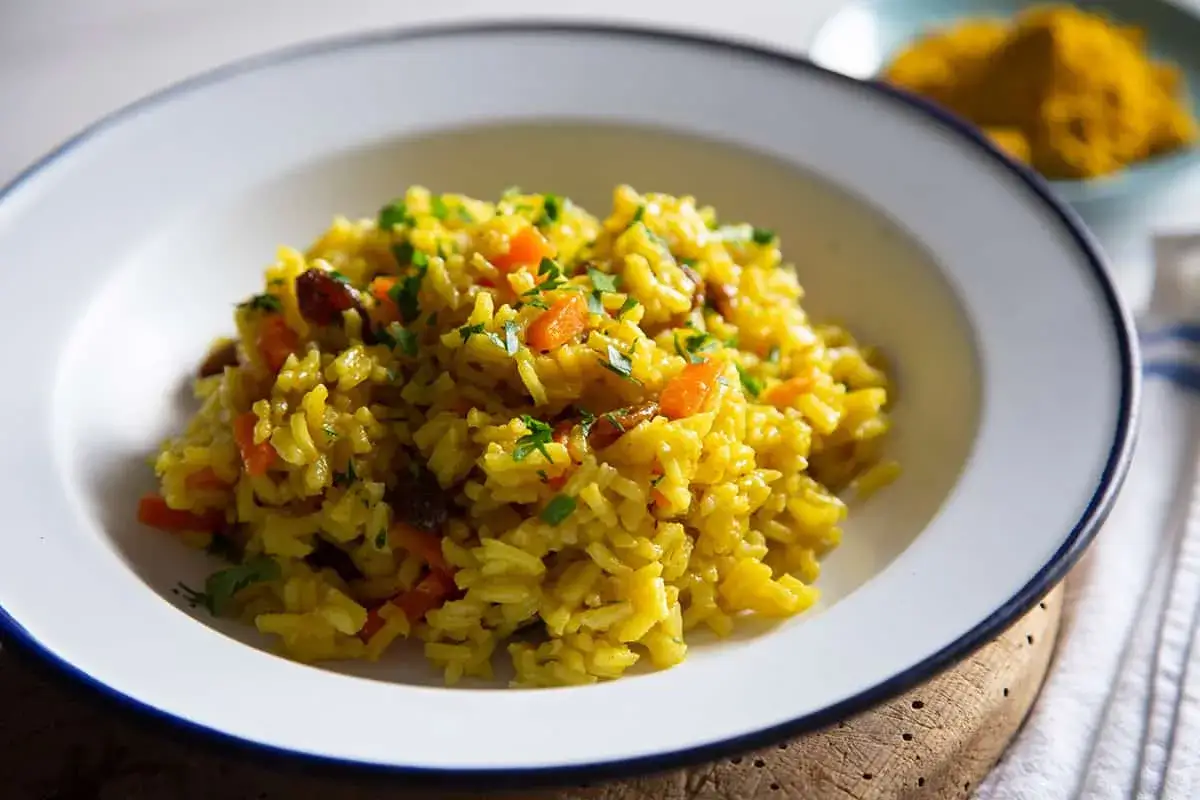 Un plato de arroz al curry con zanahoria y pasas, adornado con perejil fresco. Al fondo, un montículo de cúrcuma en polvo.