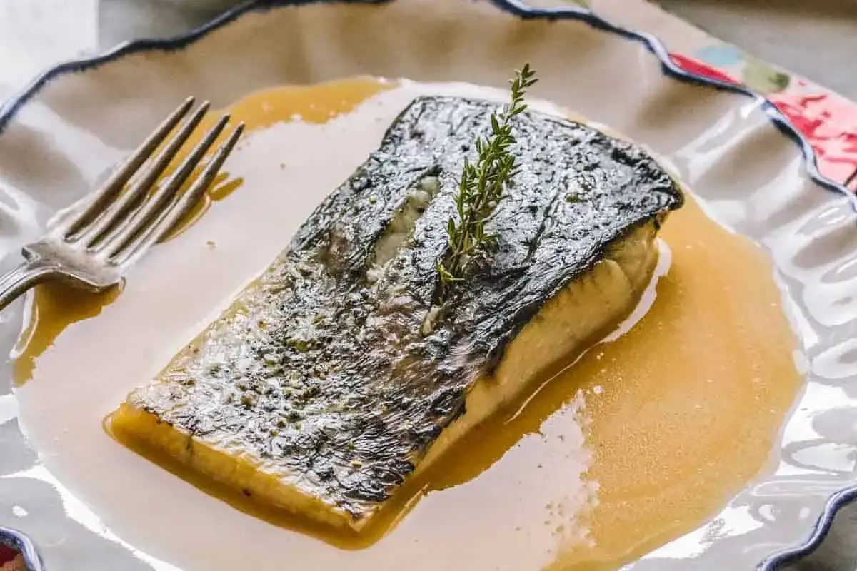 Filete de pescado crujiente ba&ntilde;ado en una cremosa salsa beurre blanc, adornado con una ramita de romero.