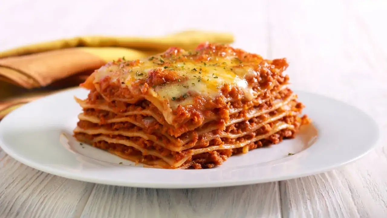 Porción generosa de lasaña boloñesa, con capas de pasta, carne y queso fundido, servida en un plato blanco.