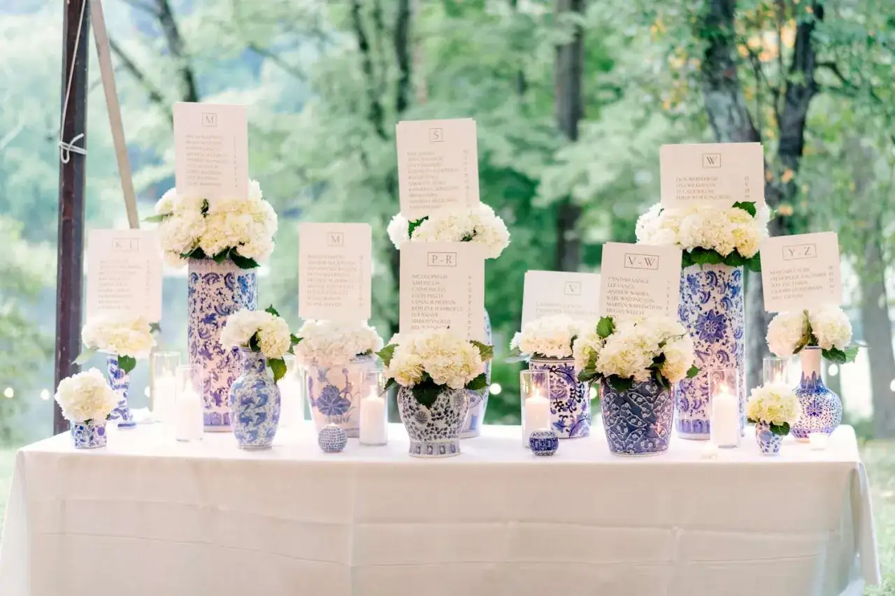 Nombres de mesas de boda con arreglos florales blancos y jarrones azules.