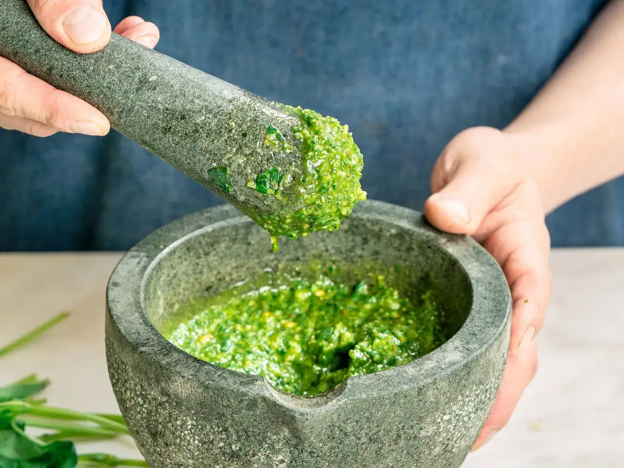 Manos moliendo albahaca fresca en un mortero para hacer una vibrante salsa pesto.