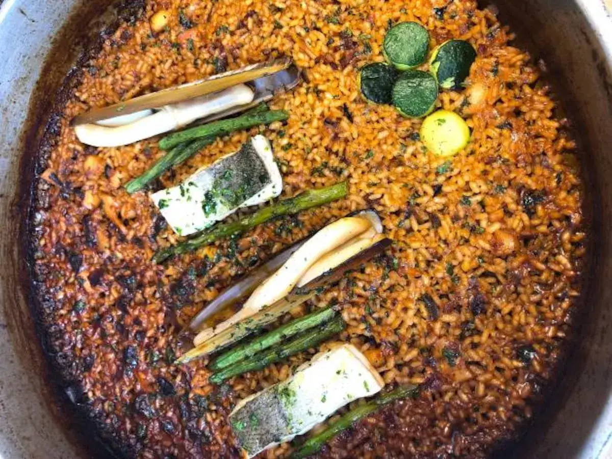 Paella quemada con trozos de pescado, espárragos y navajas. El arroz tiene un color oscuro y se ve un poco pegado al fondo.