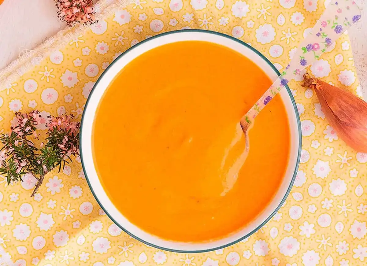 Un taz&oacute;n de cremosa salsa americana, con una cuchara floral lista para probar. Un toque de cebolla y flores decoran la mesa.