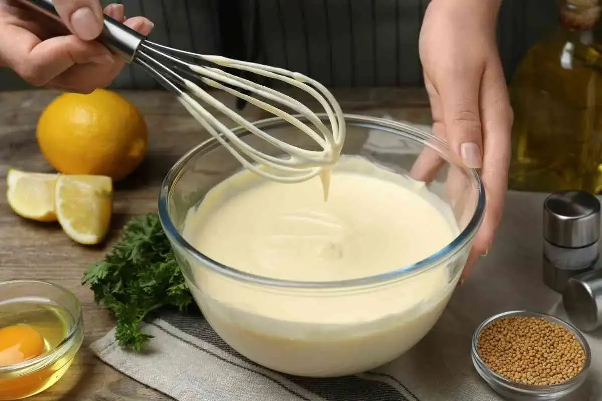 Manos batiendo para crear mayonesa casera. Ingredientes como limón, huevo y perejil alrededor.