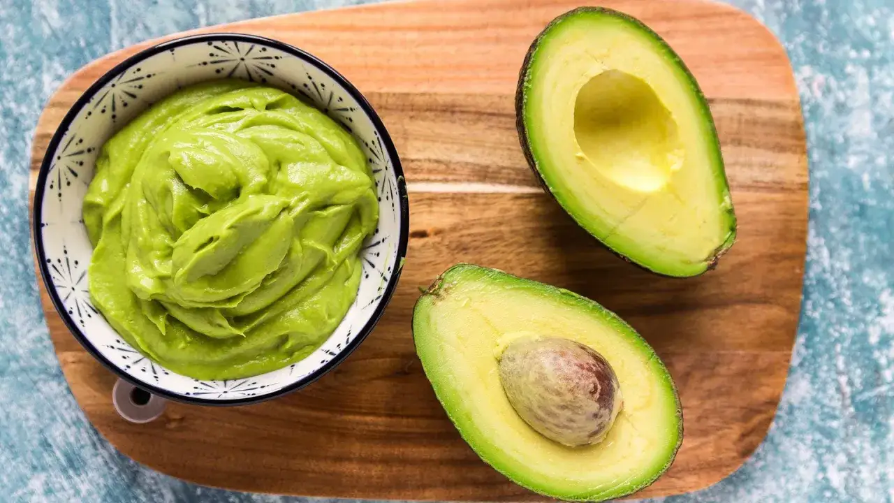 Un tazón con mayonesa de aguacate cremoso junto a dos mitades de aguacate fresco sobre una tabla de madera.