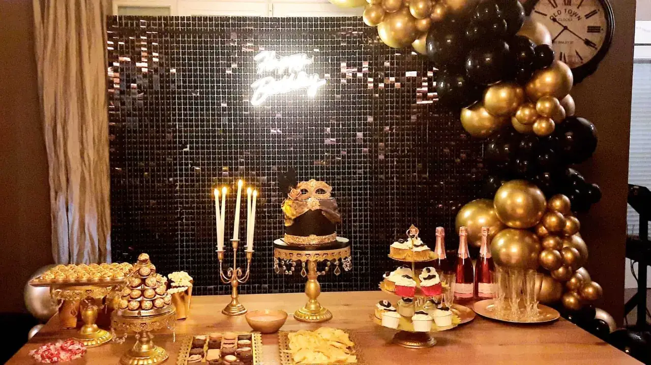 Elegantes ideas para cumplea&ntilde;os adultos en casa: mesa dulce con pastel tem&aacute;tico, cupcakes, dulces dorados y globos negros y dorados.