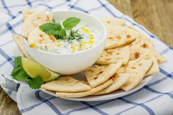 Un cuenco de cremosa salsa tzatziki, decorado con hierbas frescas y un chorrito de aceite, servido con pan de pita crujiente y rodajas de lima.